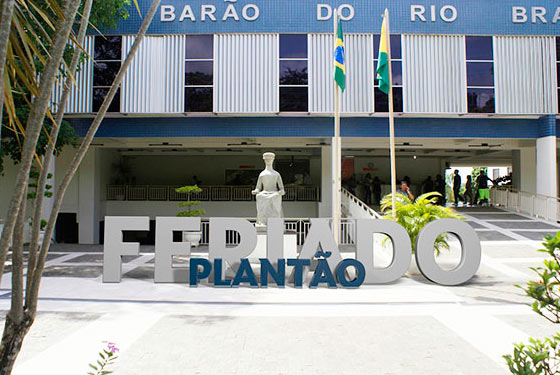 Imagem da frente do prédio do fórum Barão do Rio Branco. Na frente tem a estátua da deusa da Justiça em pedra cinza e sobreposta a palavra Feriado na cor cinza e plantão na cor azul