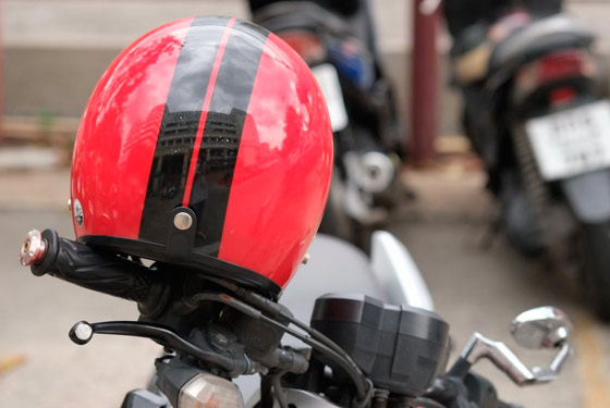 Fotografia de um capacete vermelho com duas faixas pretas verticais colocado sob o guidão de uma motocicleta.