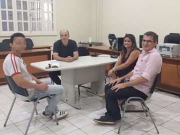 foto o juiz Gustavo Sirena em seu gabinete sentado atrás de uma mesa cinza. À esquerda um jovem com uniforme escolar, o rosto do jovem está desfocado. À direito uma mulher e um homem. Todos estão sentados e olhando para foto. Atrás tem uma bancada com uma impressora e computador sobre e encostada na parede cadeiras de escritório pretas