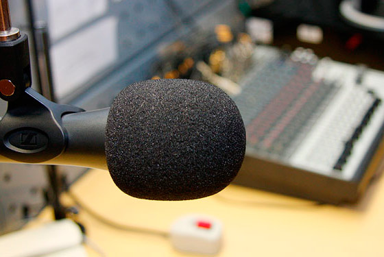 Fotografia mostrando um microfone com espuma protetora preta no primeiro plano focado e no segundo plano desfocado uma mesa de som e cabos
