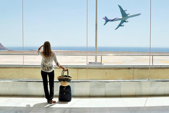fotografia de uma mulher de pé olhando por uma janela, com uma mala de rodinhas na lateral e um avião decolando no fundo