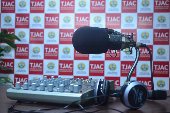 Microfone em um tripé de mesa, com fones de ouvido e mesa de som ao lado e ao fundo um banner composto por quadradinhos brancos e vermelhos com a logomarca do Tribunal dentro delas