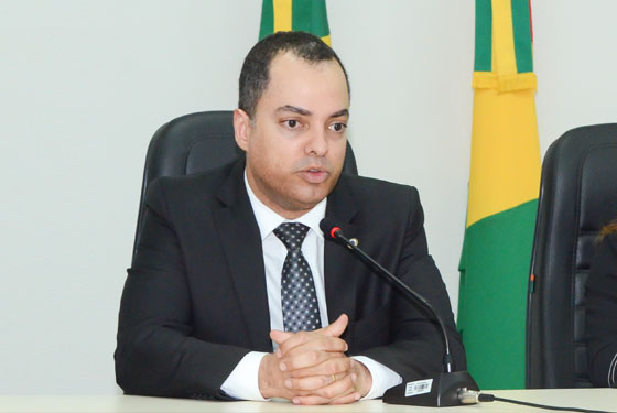 Fotografia do juiz Hugo sentado em uma mesa, com as mãos cruzadas sobre a mesa e bandeiras atrás