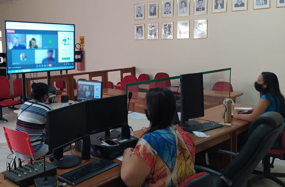 Fotografia da sala de audiência do Fórum em Tarauacá. Aparecem três mulheres sentadas na mesa em formato de T, separadas por placas de acrílico. Duas estão olhando para um monitor de TV no fundo esquerdo da imagem e outra olha para a tela do computador na frente dela.