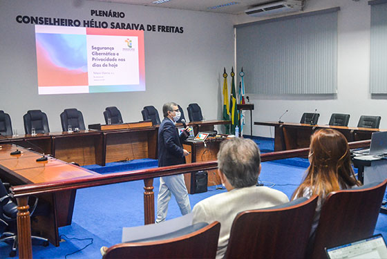 Foto do auditório do TCE, mostrando a frente com uma mesa grande em formato de U. Na parte central estão as autoridades presentes no dispositivo de honra. No primeiro plano mostra parte da plateia, com a desembargadora-presidente do TJAC sentada ao lado de um senhor.