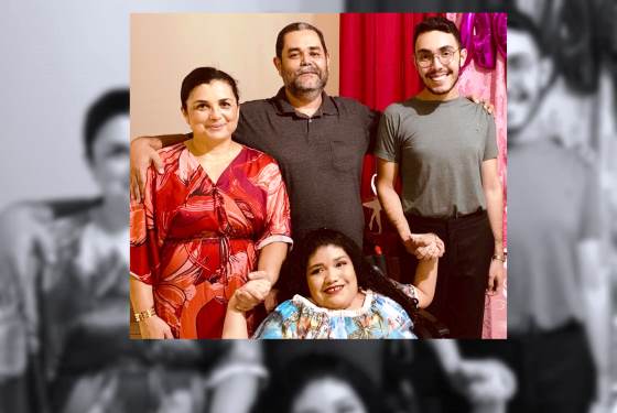 Foto da família com a mãe com um vestido vermelho no canto esquerdo da imagem, ao lado o pai, e ao lado o irmão. Na frente deles, Ana Clara Vitória, sentada em uma cadeira de rodas, segurando as mãos da mãe e do irmão. Todos olham sorridentes para a foto.