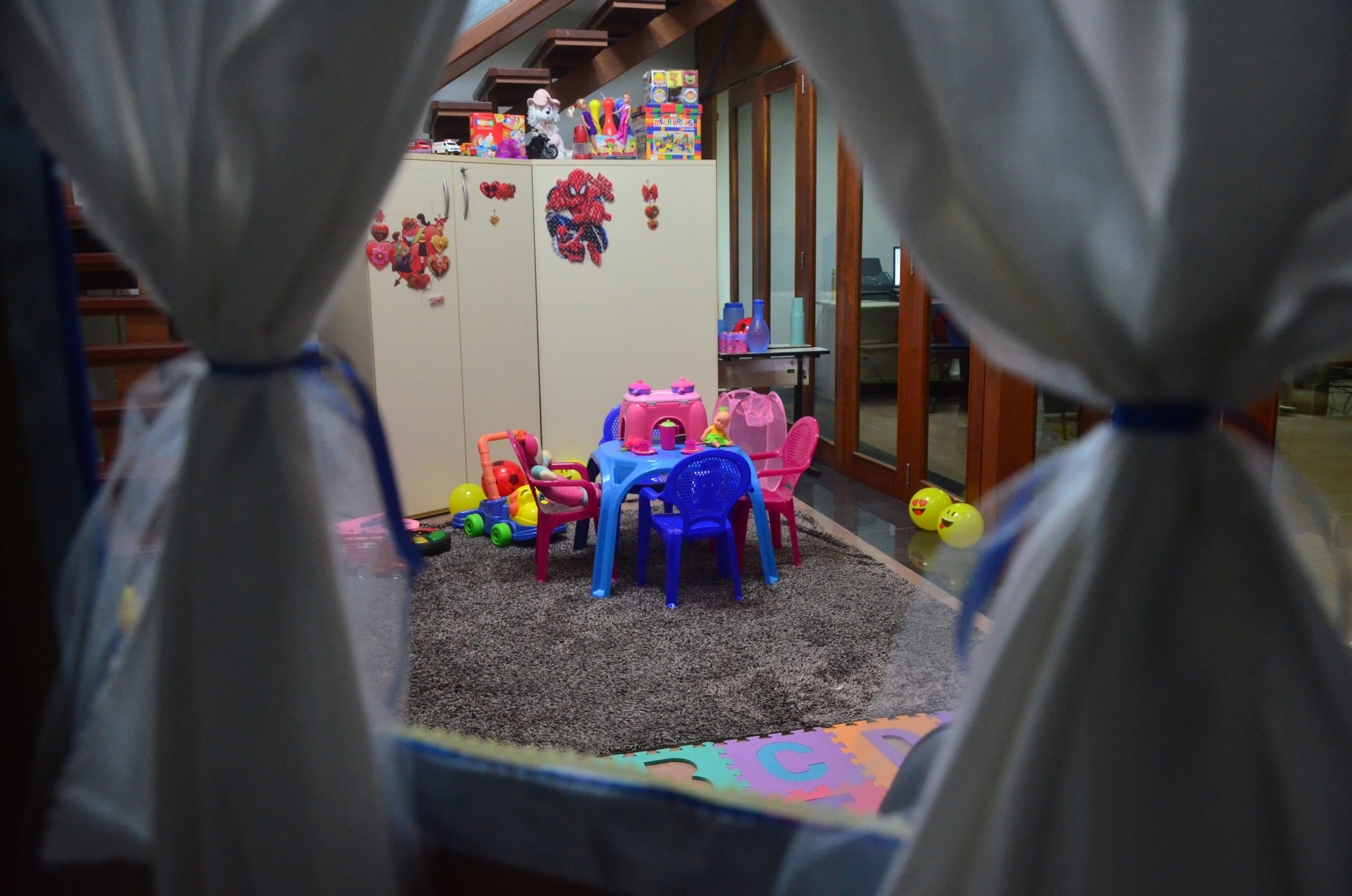 Foto da janela de uma casinha de brinquedo, mostrando mesas, armários, tapetes, o ambiente todo preparado para acolher crianças