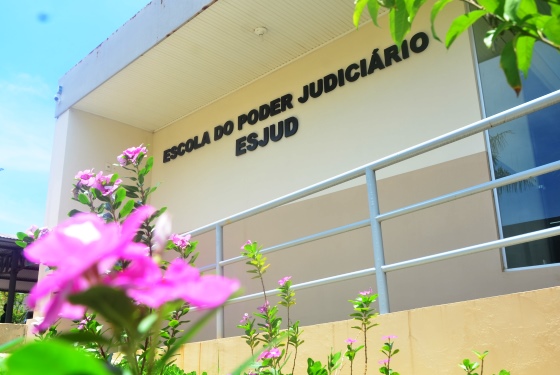 Foto da faixada da Escola do Poder Judiciário tirada de baixo para cima. Há flores roxas no primeiro e segundo plano da foto, emoldurando a parede com nome da escola pendurado