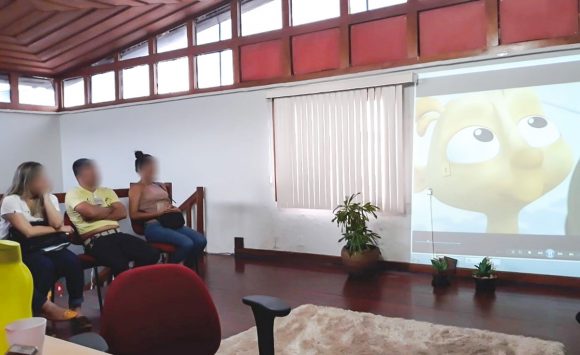 Imagem de participantes do grupo sentados à direita e a tela de projeção com um vídeo sendo exibido