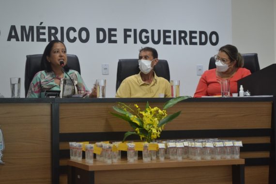 Foto da mesa de honra com a assistente social falando e duas pessoas olhando para ela