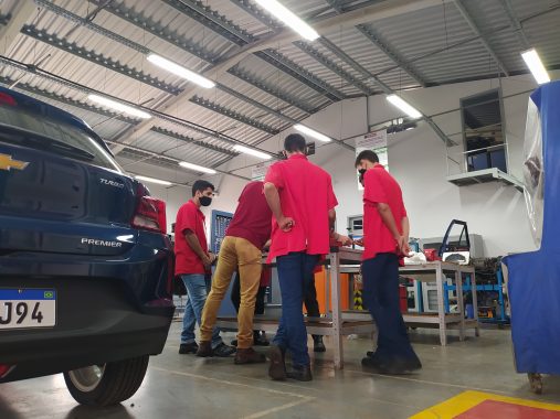 Imagem de um grupo de estudantes reunidos em volta de uma mesa com um motor de carro. Eles estão em uma oficina escola