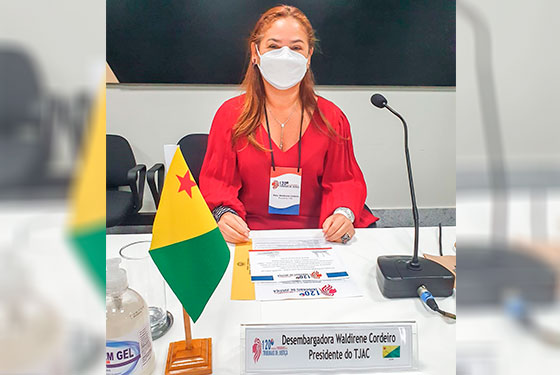 Desembargadora Waldirene Cordeiro sentada em uma mesa com a bandeira do Acre na frente