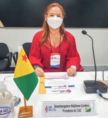 Desembargadora Waldirene Cordeiro sentada em uma mesa com a bandeira do Acre na frente