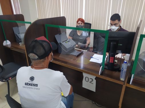 Fotografia do balcão de atendimento, onde Samuel está sentado e atrás da mesa com protetor de acrílico na frente conversam com Samuel