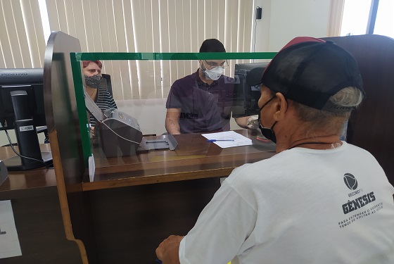 Fotografia do balcão de atendimento, onde Samuel está sentado e atrás da mesa com protetor de acrílico na frente conversam com Samuel