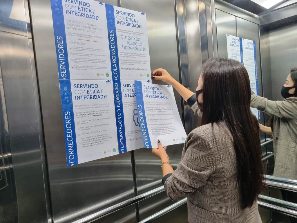 imagem de servidora grudando cartazes de conscientização sobre ética e integridade nas paredes do elevador