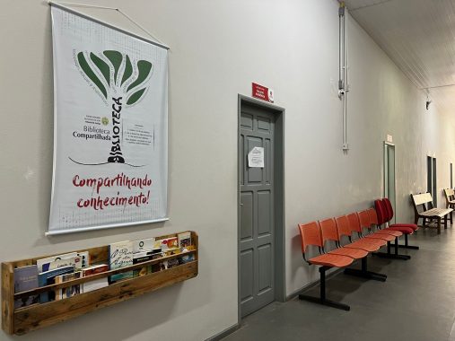 Corredor com cadeiras laranjadas. Uma porta cinza. Ao lado da porta um banner escrito Biblioteca Compartilhada e abaixo uma caixinha com livros.