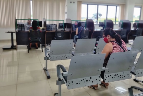 Imagem do setor de atermação dos Juizados. Na foto estão as cadeiras de espera, que são de metal e tem uma mulher sentada aguardando e na frente um balcão com várias baias de atendimento, sendo que mais duas pessoas são atendidas por servidores nessas baias