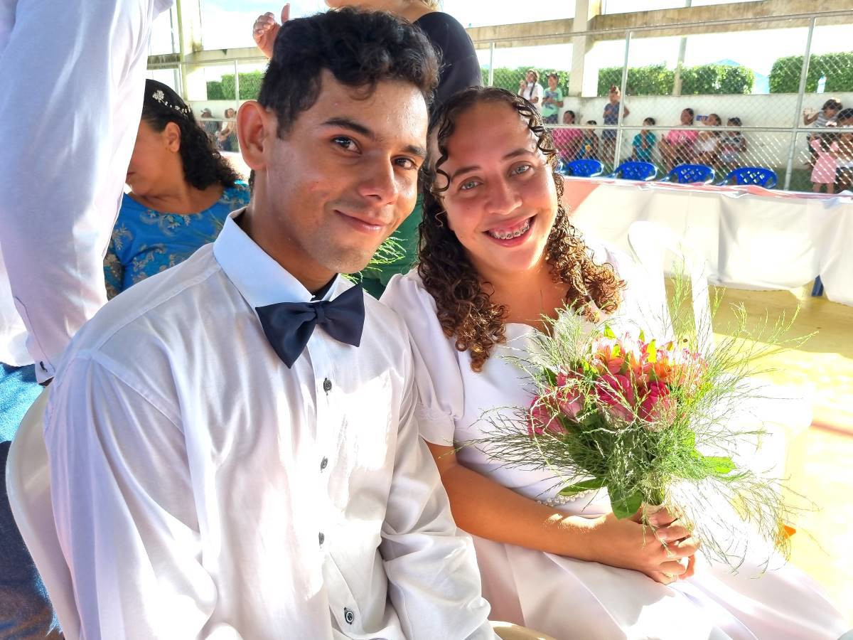 Casal de noivos posando. Mulher segurando buquê de flores. Ambos estão de branco. 