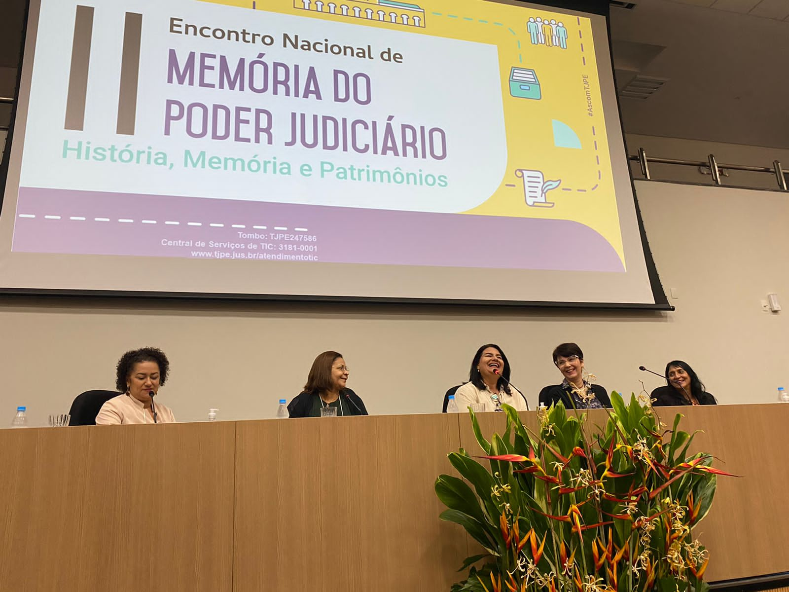 Foto mostrando a mesa com cinco expositoras sentadas atrás. Na frente tem um grande arranjo com flores laranja e verdes e atrás a tela do data show projetando o título do evento "Memória e Poder Judiciário"