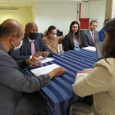 Cinco pessoas sentadas em uma mesa. Um deles olha par ao papel e o restante conversa entre si.