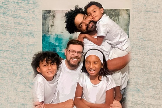Foto do casal, sendo um homem branco e outro negro. Um dos pais está com o filho no colo e um menino e a menina na frente do outro pai