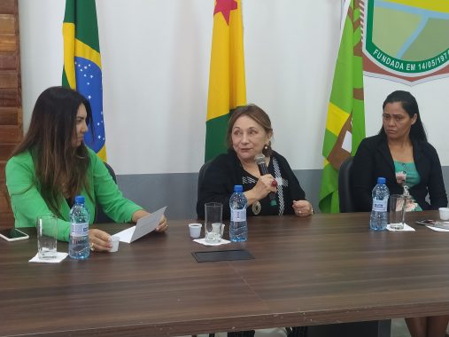 Desembargadora Eva ao microfone falando e a juíza Andréa e a vereadora de Assis Brasil olham para ela