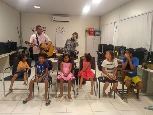 Seis crianças sentadas, duas olhando para a pessoa que está tocando violão. O ensaio ocorre em uma sala que funciona como laboratório de informática, no Educandário Santa Margarida.