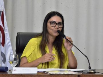 A juíza Andrea Brito está vestida de amarelo falando em um microfone que está na mesa a sua frente