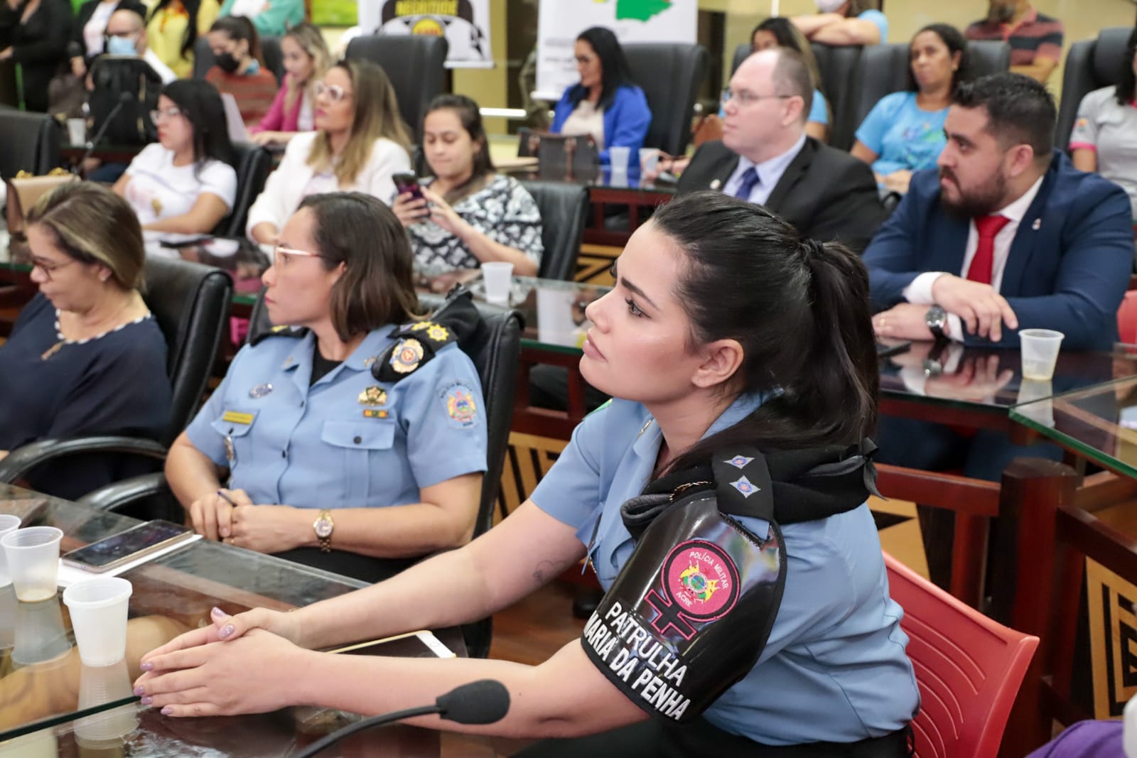 imagem de uma policial da patrulha maria da penha sentanda olhando para frente ao redor várias pessoas sentadas