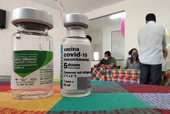 Foto dos frascos da vacina contra influenza e covid no primeiro plano focado e ao fundo imagem de uma mesa onde uma pessoa está passando suas informações