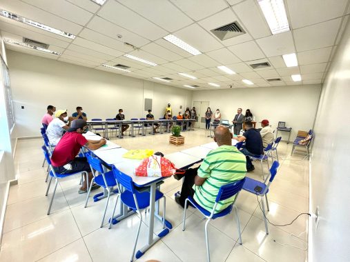 Sala com carteiras escolares em formato de U e homens e mulheres sentados olhando a parede ao fundo, onde estão a equipe coordenadora de pé