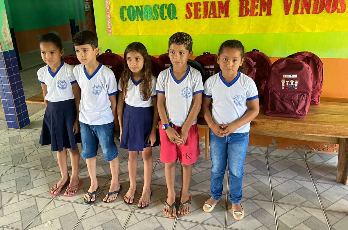 Foto de cinco crianças com uniformes escolares de pé alinhadas uma do lado da outra na frente das mochilas dos kits escolares
