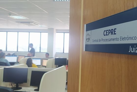 Foto da porta entreaberta com a placa da Cepre e mostrando as baias com computadores e pessoas trabalhando