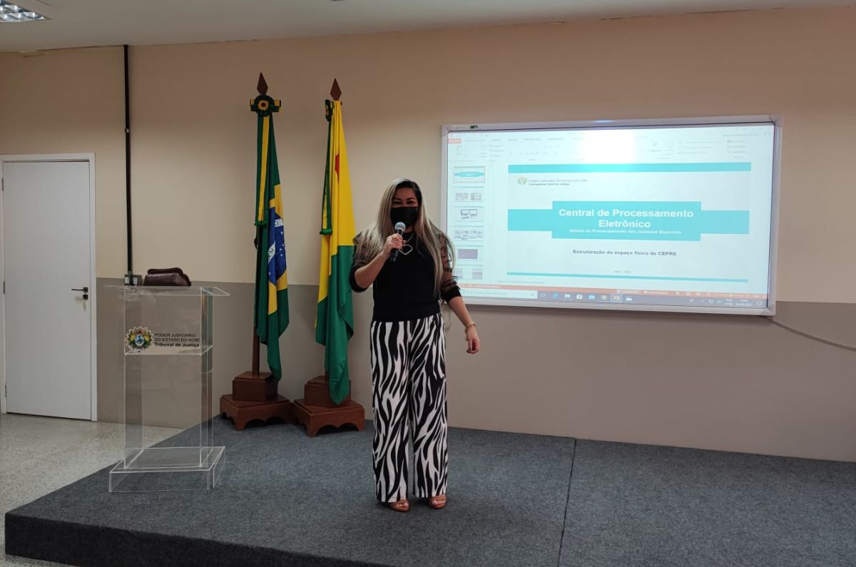 Foto da psicologa de pé falando ao microfone, atrás dela um quadro com projeto de slides e das bandeiras do Brasil e do Acre no lado esquerdo