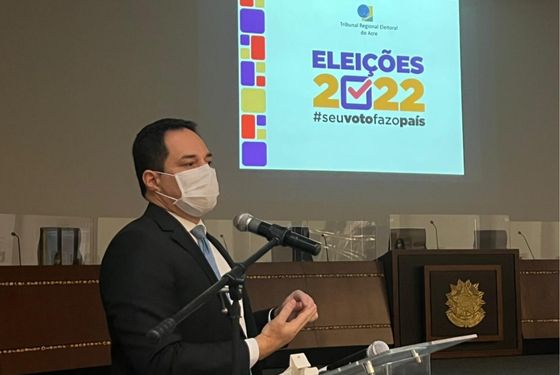 Foto do juiz eleitoral falando no púlpito e atrás aparece um slide projetado na parede com a logomarca do TRE e o texto "Eleições 2022. #seuvotofazopaís