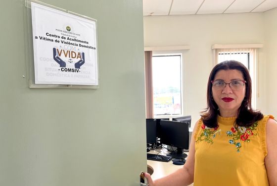 Psicóloga Eunice Guerra segura a porta da sala de atendimento aberta. Ela esta com uma blusa amarela com bordados de flores e usa batom vermelho. Na porta bem uma placa com logomarca do programa Comv-vida e do TJAC e está escrito "Centro de Acolhimento a Vítima de Violência Doméstica"