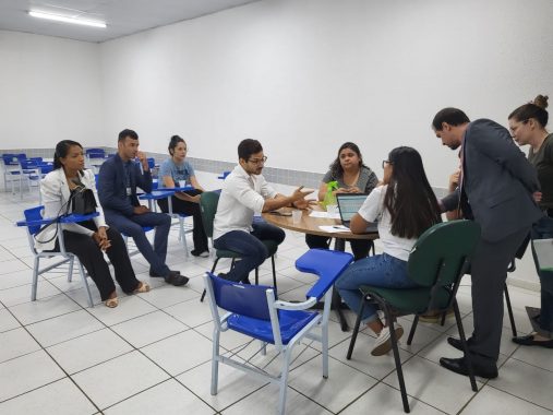 Sala de aula com uma mesa em que está as partes de uma audiência de conciliação e três alunos de Direito assistindo os atos processuais