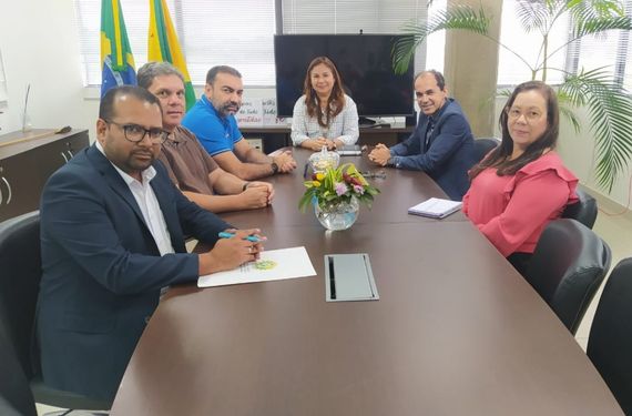 Presidente vestida de branco. Sentada na cadeira. Cinco pessoas estão ao redor da mesa sentadas