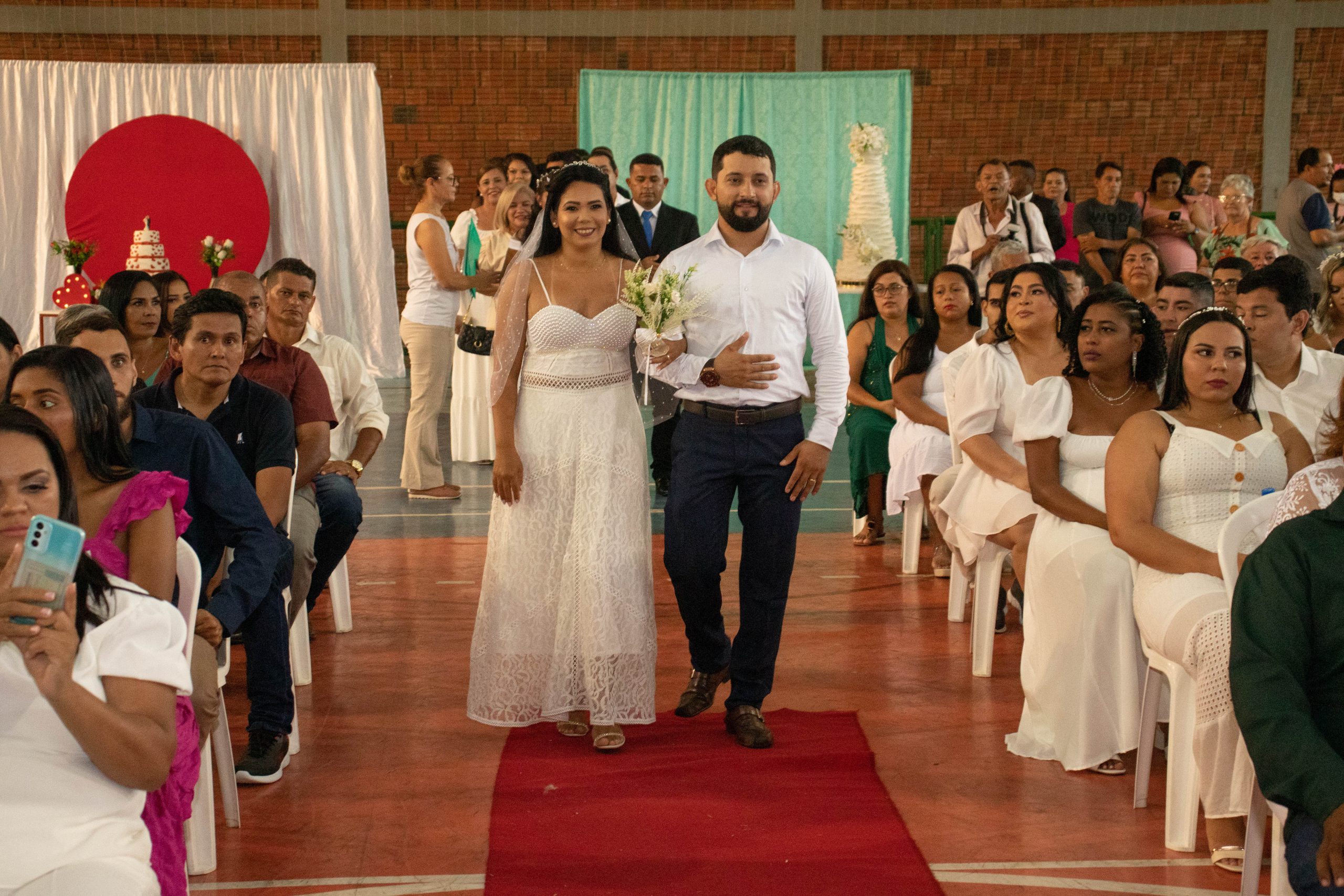 Os noivos entram sorrindo num tapete vermelho enquanto os outros noivos sentados assistem a cerimônia