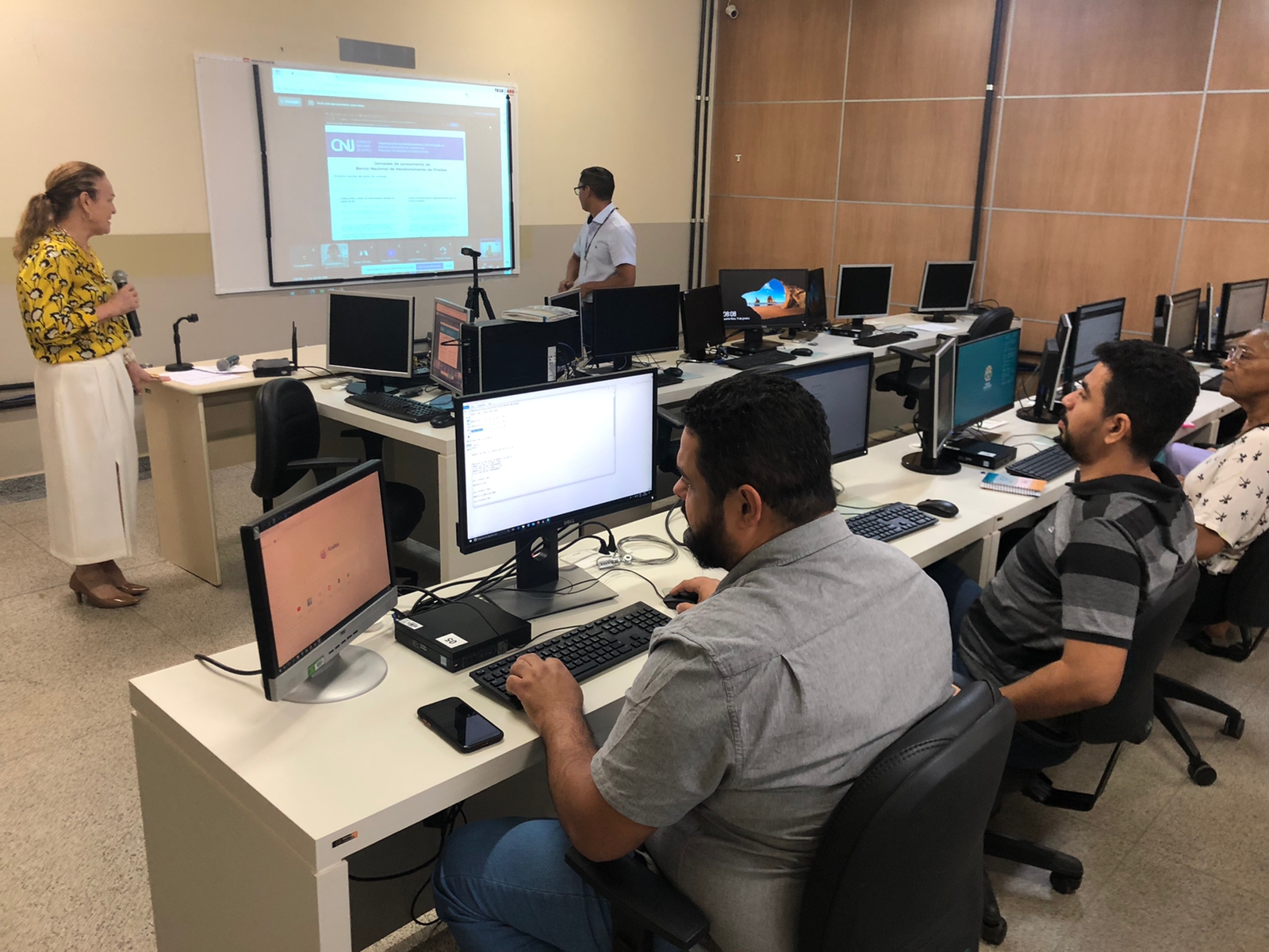 Foto da oficina do BNMP na Esjud. Há uma projeção da ferramenta na tela. A desembargadora Regina Ferrari, diretora da Escola, está ao microfone. O servidor Josemar auxilia na apresentação. Os alunos são servidores que estão no laboratório de informática, tendo o aprendizado diretamente no computador.