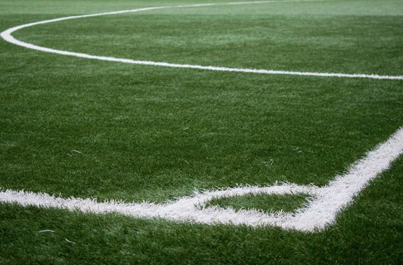 foto de um canto de campo de futebol. A grama esta verde, tem linhas brancas traçadas, mostrando o local de escanteio e uma parte da grande área