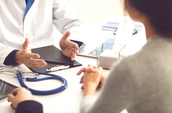 foto de um médico atendendo uma mulher. Mostra-se o médico do pescoço para baixo até a cintura. O médico está de jaleco branco com as mãos sobre a mesa. Na mesa tem estetoscópio, prancheta, notebook, lápis e uma caixinha com papeis brancos. A mulher esta de costas segurando as mãos juntas