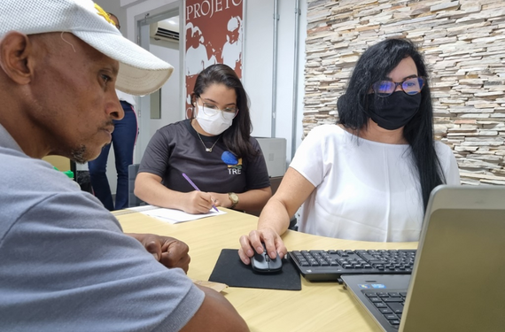 Imagem de duas mulheres atendendo uma pessoa em situação de rua. É um homem, com pele negra e usa bone, tem barba e bigode ralo, com fios brancos