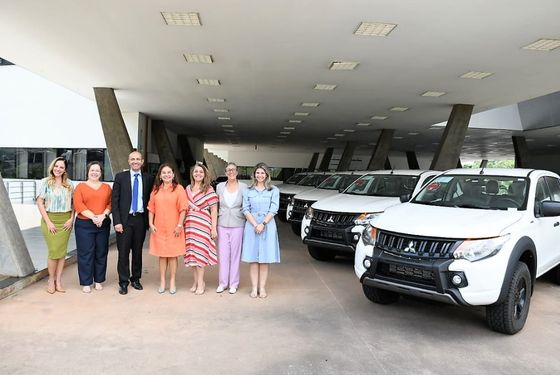 Na direita uma fileira de caminhonetes brancas estacionadas na diagonal. Na esquerda seis mulheres e um homem estão em posicionados lado a lado posando para foto.