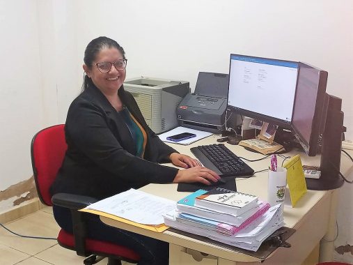 Foto de servidora sentada na mesa de trabalho sorrindo. Ela tem cabelos escuros e presos, usa óculos, e um blazer preto sob uma blusa listrada
