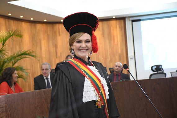 Desembargadora Regina olhando para câmara e sorrindo. Ela veste toga presta com detalhes vermelhos, capelo, tem uma faixa atravessada na diagonal sobre a toga e também usa o grão-colar. Ela é branca, tem cabelo longo, loiro e preso lateralmente