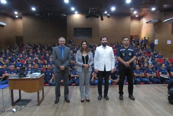 Foto das autoridades de pé no palco, ao fundo a plateia sentada e lotada