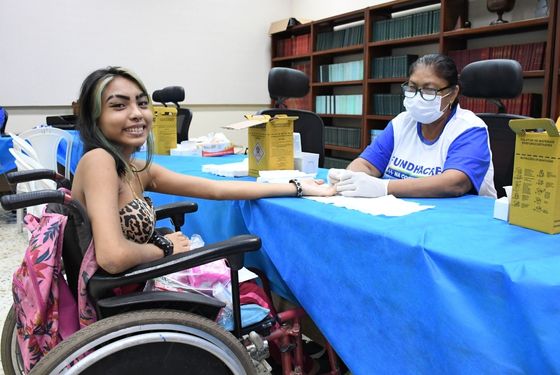 foto de uma jovem indígena cadeirante sendo atendida com serviço de saúde. A jovem tem mexas loiras no cabelo liso, preto e de cumprimento mediado. Veste um tomara que caia com estampa de onça e está sorrindo