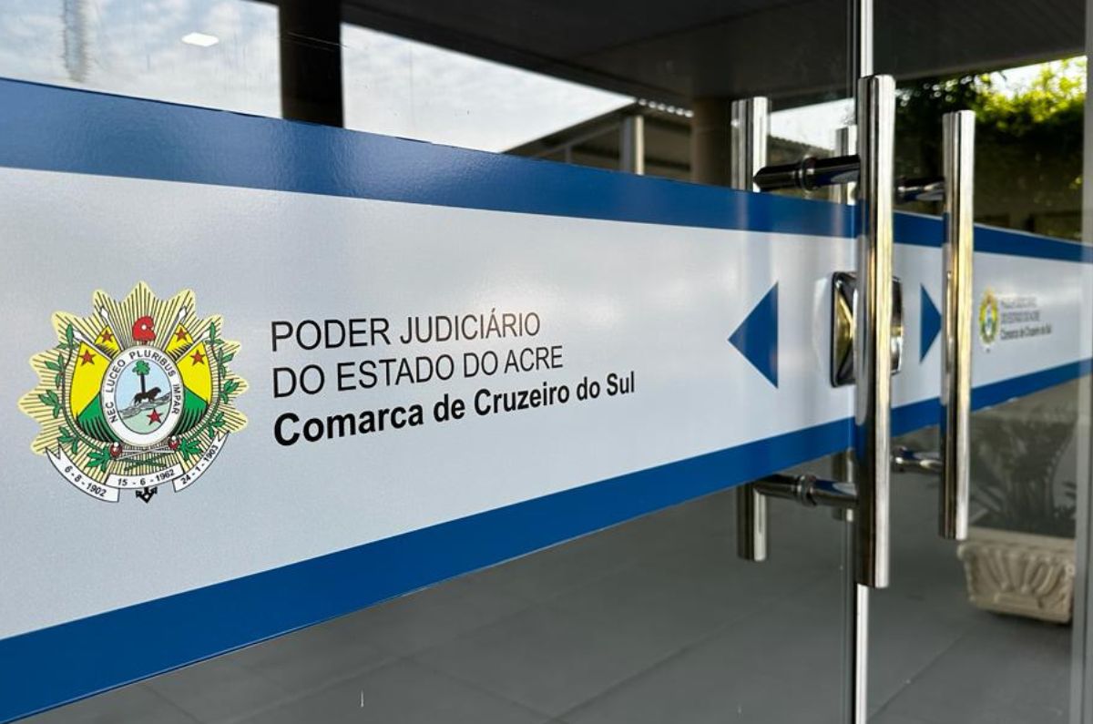 Foto da porta de entrada do Fórum de Cruzeiro do Sul. É uma porta de vidro com adesivo em tons azuis com logomarca do Estado e escrito "Poder Judiciário do Estado do Acre: Cruzeiro do Sul". No primeiro plano desfocada há flores rosa e verde.